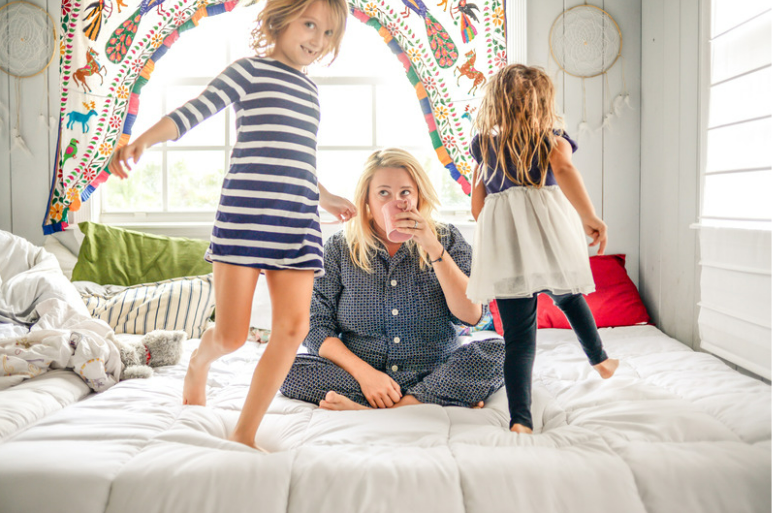 Een moeder zittend op een bed terwijl de kinderen spelen. Een voorbeeld van adaptief ouderschap.